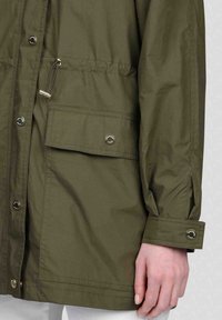 Veste légère vert olive avec fermeture éclair, boutons pression, cordon de serrage ajustable et une poche latérale. Présente une finition légèrement texturée.