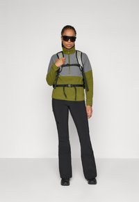 Fleece bunda so zelenými a šedými panelmi, s vysokým golierom a polovičným zipsom. Čierne nohavice so priliehavým strihom a outdoorové topánky.