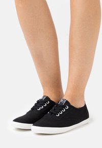 Svarta canvas sneakers med vit gummisula och öglor, med svarta snören och en logotagg på plösen. Platt design och lågskuren.