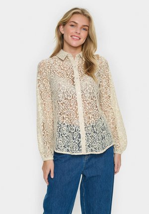 Blouse en dentelle beige avec des manches longues, un devant à boutons et un motif floral. Portée sur un jean en denim légèrement ample.