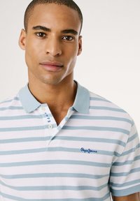 Pruhovaná polokošela v svetlom modrom a bielom prevedení, z bavlneného materiálu, s modrým logom na hrudi a trojcovou gombíkovou légou so kontrastnými gombíkmi.