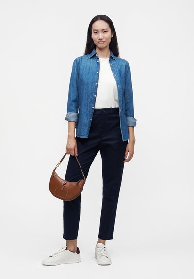 Femme debout avec de longs cheveux noirs portant une chemise en jean bleue, un t-shirt blanc, un pantalon noir, des baskets blanches et tenant un sac à main marron.