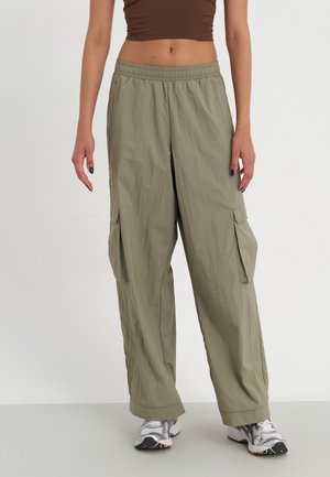 Persoon die losse olijfkleurige cargobroek draagt met een elastische tailleband, witte sportschoenen en een bruin cropped topje, staand tegen een effen achtergrond.