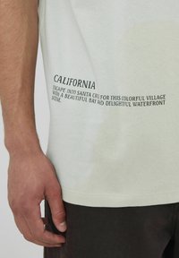 T-shirt en coton vert clair avec un col rond, des manches courtes et un design imprimé sur le côté présentant un texte inspiré de la Californie.
