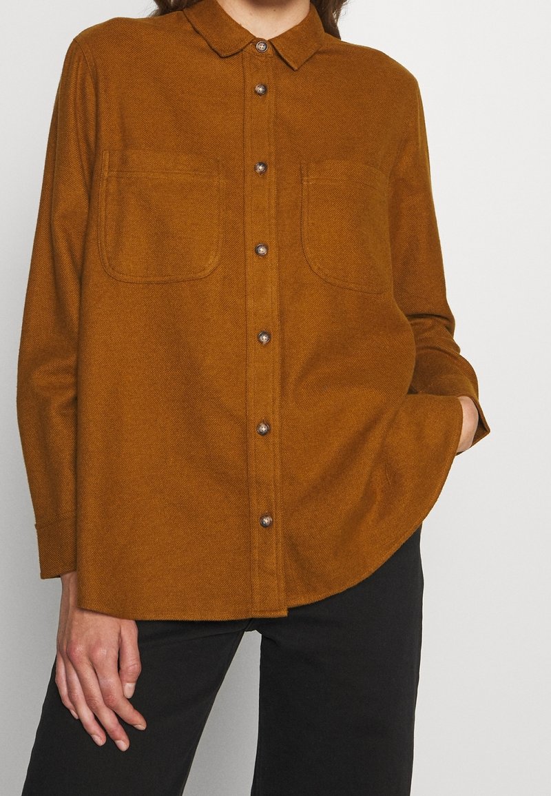 Chemise marron boutonnée avec col et poches poitrine portée par une personne avec une main dans la poche et un pantalon noir visible.