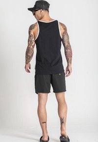 Čierny top bez rukávov s bielym lemovaním, spárovaný s tmavými šortkami. Obsahuje tetovania na ramenách a nohách a čiapku s sieťovinou vzadu. Nosené sú šľapky.