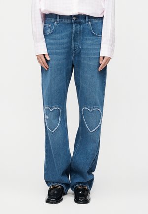 Osoba ubrana w niebieskie dżinsy z postrzępionymi, w kształcie serca łatkami na kolanach, jasnoróżową koszulę i czarne mokasyny ze złotymi detalami.