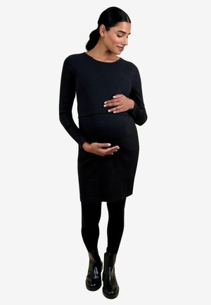 Schwangere Frau mit dunklem Haar in einem schwarzen Langarmkleid, schwarzen Leggings und schwarzen Stiefeln, die sanft ihren Bauch hält und vor einem weißen Hintergrund steht.