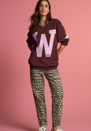 Maroon stor sweatshirt med stor vit "W" logotyp och texten "WICKED"; ihop med beige leopardmönstrade byxor och vita sneakers. Pink bakgrund.
