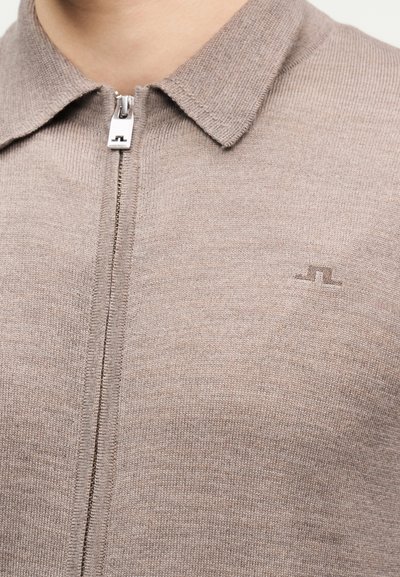 Κοντινή λήψη ενός μπεζ πλεκτού πουλόβερ με φερμουάρ, γιακά και μικρό κεντημένο λογότυπο στο αριστερό στήθος.