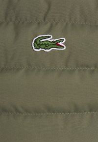 Zielone haftowane logo krokodyla z czerwonym otworem ust na oliwkowozielonej pikowanej tkaninie z poziomymi liniami przeszyć.