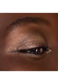 Primer plano de un ojo marrón cerrado con largas pestañas, ceja oscura y sombras de ojos brillantes en tonos dorados y bronce.