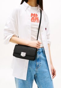 Donna che indossa jeans chiari, top corto bianco con grafica, camicia bianca aperta, mentre tiene una borsa a tracolla nera con chiusura argentata.