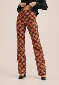 Pantalons à motifs dans un design floral vibrant en rose, orange et noir ; forme évasée ; tissu léger ; portés avec un haut noir et des talons.