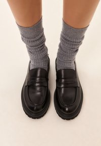 Černé kožené mokasíny s hladkým povrchem a polstrovanou podrážkou, doplněné šedými žebrovanými ponožkami sahajícími do poloviny lýtek.