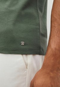 Vihreä teksturoitu polo-paita, jossa on hunajakennokuvio, ja pienen hopeisen logon kanssa helman lähellä. Yhdistetty vaaleanvärisiin housuihin.