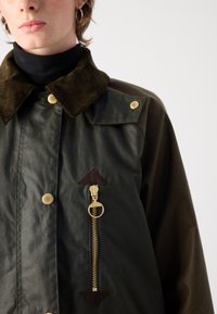Olijfgroene jas met een corduroy kraag, gouden hardware en een ritszak met een bruine driehoekige accent. Textuur van de stof is duidelijk zichtbaar.