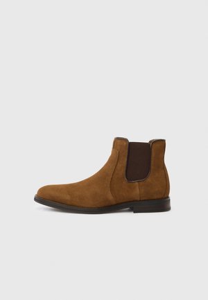 Brauner Chelsea-Stiefel aus Wildleder mit dunklen elastischen Seitenpartien und einer schwarzen Sohle, dargestellt in einer linken Seitenansicht vor einem schlichten Hintergrund.
