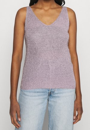 Kobieta w bezrękawniku wykonanym z lawendowego dzianiny z dekoltem w serek, zestawionym z jasnoniebieskimi dżinsami, stojąca na prostym jasnym tle.