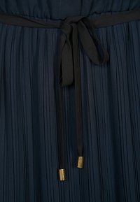 Veckat mörkblått tyg med ett knutet svart tygskärp som har guldfärgade metall-ändhylsor.