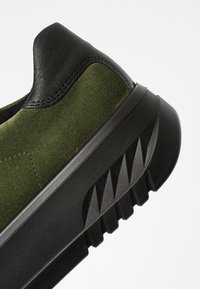Green suede sneaker with black leather accents. Features a textured rubber sole with angled patterns for grip and a cushioned heel.