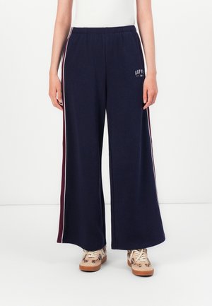 Pantalon bleu marine ample avec bandes blanches et bordeaux sur les côtés, taille élastique, logo « GAP » sur la cuisse, porté avec des baskets beiges et un haut blanc.