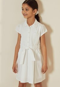 Robe en coton blanc à manches courtes, col, bouton sur le devant et ceinture à la taille. Présente un motif texturé et une petite poche sur la poitrine.