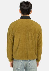 Khaki fleece jas met een gestructureerde, zachte afwerking, voorzien van een zwarte kraag en een ontspannen pasvorm, gezien vanaf de achterkant.