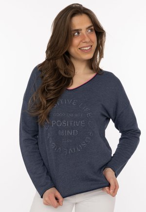 Frau mit langen braunen Haaren lächelt, während sie einen dunkelblauen Pullover mit dem Text „Positive Mind“ trägt, kombiniert mit weißen Hosen.