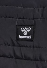 Svart tyg med en slät, puffig textur har en rektangulär svart lapp som visar den vita "hummel"-logotypen ovanför ett stiliserat emblem.