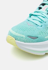 Chaussure de course légère avec une tige en mesh turquoise, finition texturée, semelle intermédiaire blanche et semelle extérieure jaune, présentant des accents subtils en gris et en rose.