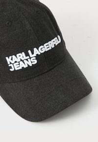 Juodos džinsinės kepurės su balta siuvinėta užrašu: "KARL LAGERFELD JEANS." Dizainas apima kreivą kraštą ir tekstūruotą audinį.