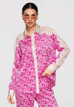 Femme portant une chemise et un pantalon à motifs floraux rose vif avec des manches et un col en dentelle, posant avec des lunettes de soleil teintées orange.
