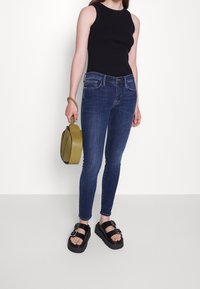 Jeans de denim azul ajustados, top negro sin mangas, sandalias negras gruesas y un bolso redondo verde olivo. Textura plana con detalles mínimos.