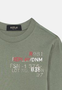 Grünes Baumwoll-T-Shirt mit langen Ärmeln, das das Logo "REPLAY" in roten und weißen Schriftzügen zeigt, ergänzt durch zusätzliche numerische Details in Grau.