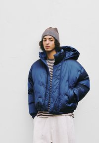 Puchowa kurtka w głębokim granatowym kolorze z błyszczącą fakturą, z kapturem, zapięciem na guziki i elastycznymi mankietami, noszona na paskowanym topie i jasnych spodniach.