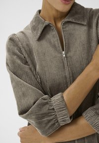 Giacca zip-up in corduroy color taupe, con colletto, texture a coste e maniche voluminose con polsini arricciati.