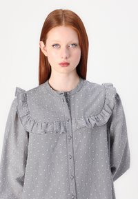 Blusa grigia a scacchi con colletto ruffle e chiusura con bottoni, caratterizzata da motivi a pois e maniche lunghe, realizzata in tessuto leggero.