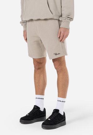 Homme portant un short beige avec le logo "Pegador", un sweat à capuche beige assorti, des chaussettes blanches avec du texte et des baskets noires, debout sur fond blanc.