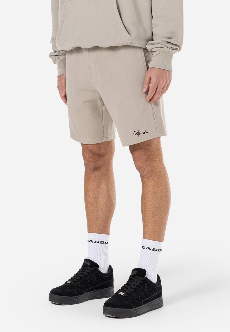 Homme portant un short beige avec le logo "Pegador", un sweat à capuche beige assorti, des chaussettes blanches avec du texte et des baskets noires, debout sur fond blanc.