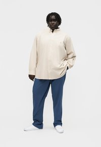 Osoba tamne puti i dredovima, odjevena u preveliku bež košulju na kockice, plave hlače i bijele tenisice, stoji na jednoličnoj bijeloj pozadini.