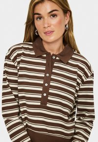 Jeune femme portant une chemise polo à rayures horizontales marron et crème à manches longues avec une patte de boutonnage et des boucles d'oreilles en argent, sur fond blanc.