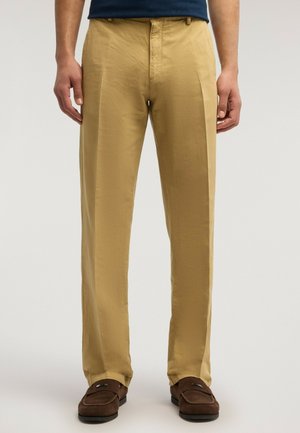 Uomo che indossa pantaloni chino beige e mocassini in camoscio marrone, in piedi contro uno sfondo chiaro e uniforme.