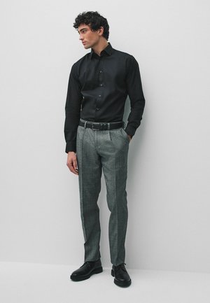 Mann trägt schwarzes Hemd mit Knöpfen, graue maßgeschneiderte Hose, schwarzer Gürtel und schwarze Schuhe, steht vor einer schlichten hellgrauen Wand.