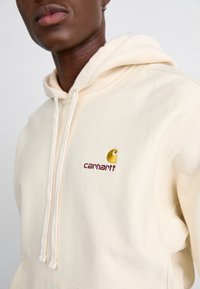 Hoodie beige chiaro realizzato in tessuto morbido, con tasca frontale, cappuccio con coulisse e logo Carhartt ricamato in oro e borgogna.