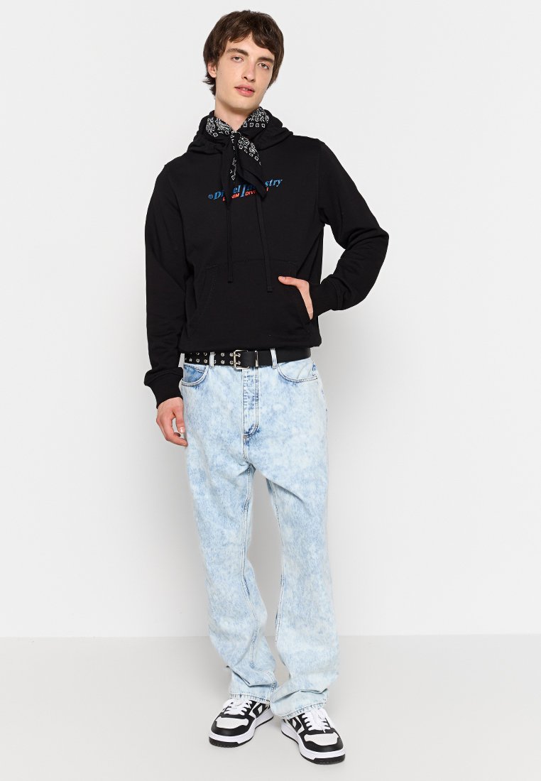 Hoodie noir avec poche avant, portant un texte bleu et rouge. Jean baggy bleu clair délavé et baskets noires avec des accents blancs.