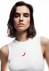Jonge vrouw met kort donker haar draagt een witte mouwloze top met een kleine rode peper geborduurd op de borst.