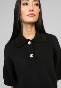 Polo shirt noir en tricot à manches courtes, présentant trois embellissements en forme de fleur de couleur blanche comme boutons et un design de col classique.