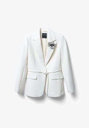 Blazer blanc sur mesure avec un col à revers, une taille ceinturée et des poches avant. Il présente un broche florale noire du côté gauche.