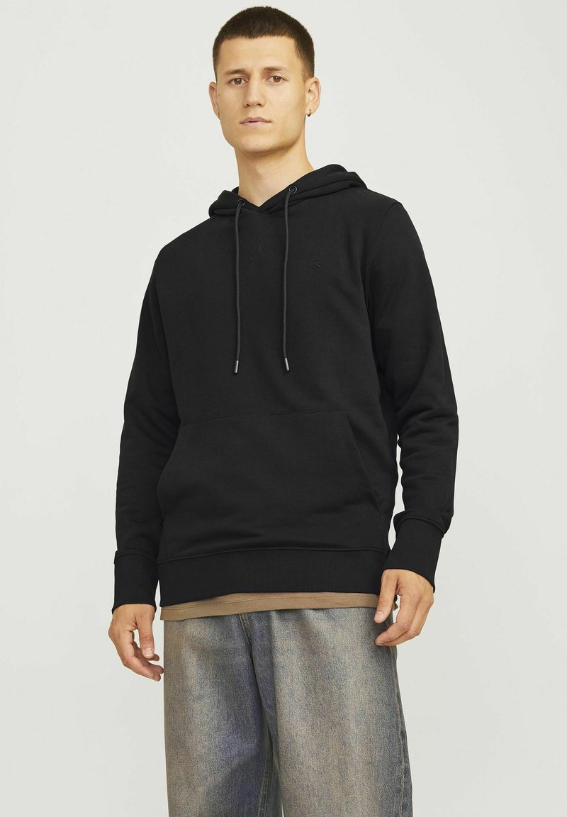 Black hoodie made of soft fabric with a kangaroo pocket, adjustable drawstrings, and ribbed cuffs and hem. Worn over light-colored pants.